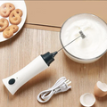 White handheld milk frother with black top on a wooden surface next to a bowl of whipped cream and cookies.
